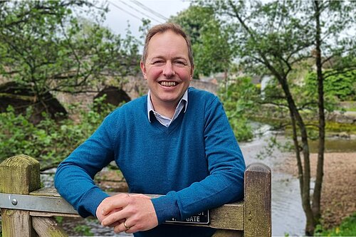 Richard Foord standing leaning on a wooden gate. He is facing the camera and smiling. He is wearing a blue jumper. Behind him are an old stone bridge, a stream, and some trees.
