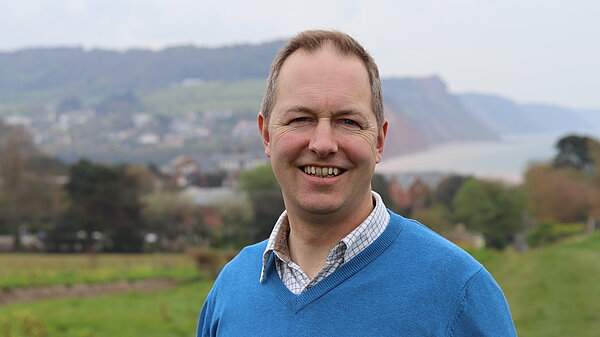 Richard Foord stood in a field with the East Devon coastline in the background