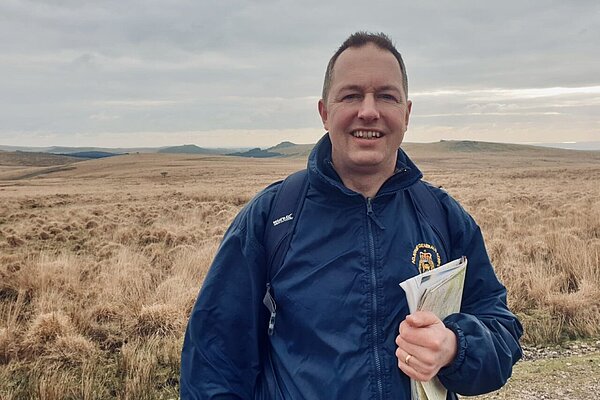 Richard Foord standing on dartmoor in a waterproof jacket and with a backpack. He is holding a map with his left arm over his chest