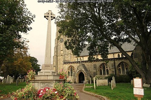 Sidmouth War Memorial in front of the Parish Church of St. Giles and St. Peter. There are wreathes and flowers laid around the memorial, with trees between it and the church building. The image also shows a path between the memorial and the doorway to the church.