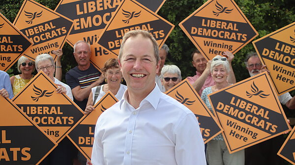 Richard Foord stood smiling in front of a crowd of supporters, who are holding Lib Dem diamonds