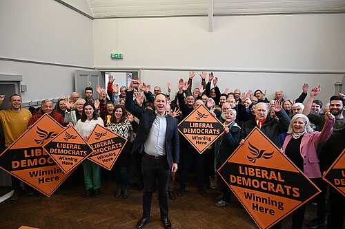 Richard Foord standing in a hall with a large group of activists holding signs reading "Liberal Democrats winning here"